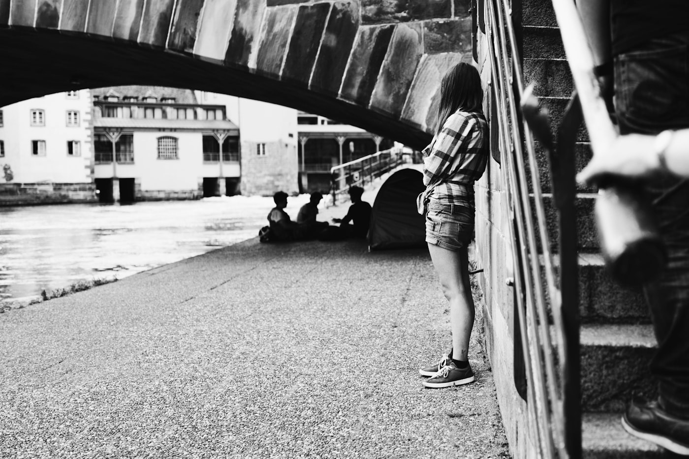 A figure leaning against a wall beneath a stone bridge by a river
